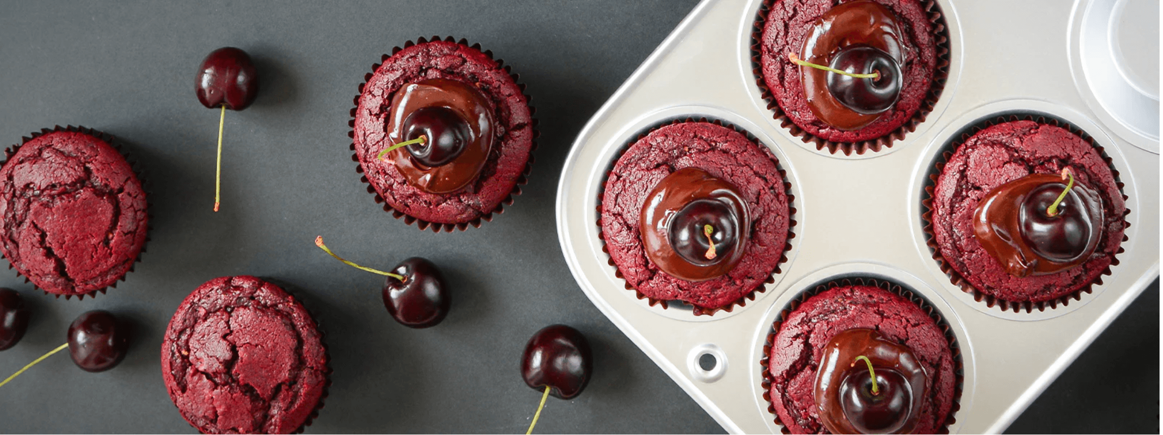 Overview of some red velvet cupcakes sitting in a Silverwood Bakeware muffin tin with chocolate icing and a fresh cherry on top. Iced and non-iced cupcakes sit on a dark grey bench beside it with strategically scattered cherries around. 